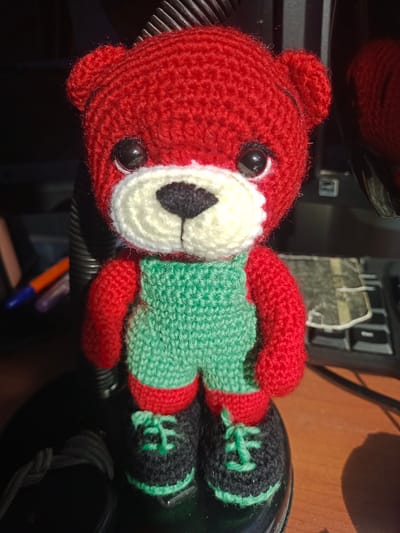 A red crocheted teddy bear wearing green overalls and black shoes with green laces stands on a dark, shiny surface. The bear has black button eyes, a white muzzle with a black nose and mouth. The background is blurred, showing parts of a keyboard, a ribbed black object, and some writing instruments. The lighting suggests the photo was taken indoors with a strong light source, creating shadows. The overall scene appears to be a desk or workspace.