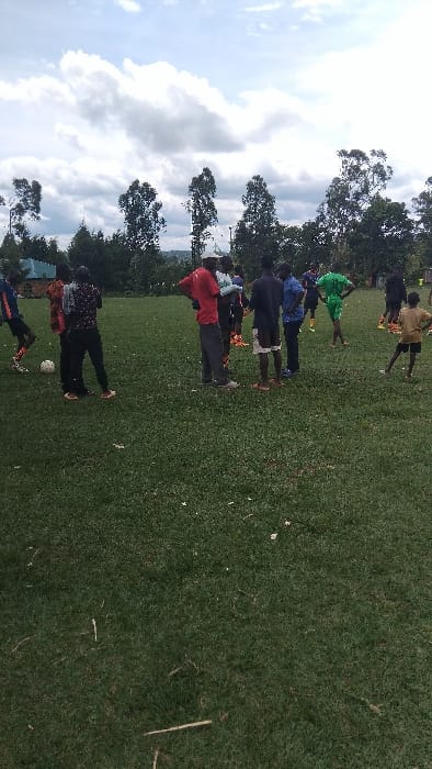The image shows a group of people on a grassy field, likely a soccer field, in Bungoma, Kenya, located at latitude: 0.553, longitude: 34.773. The scene is dominated by the verdant field and a partly cloudy sky. There are approximately 10 people on the field. Most of them are men and appear to be watching or coaching a game. A white soccer ball lies on the field in the foreground. Some men are wearing casual attire, while others are dressed in athletic clothing.  They are standing, talking, and observing, interacting casually amongst themselves. Trees line the edge of the field.