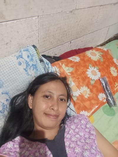 In Baron, Indonesia, a woman is lying on her side, facing the camera, with her head and upper body visible. She has dark hair and is wearing a purple top with a floral pattern. She is on a bed covered with blankets and pillows. One pillow in the background has a large orange floral pattern, and another to her left has a blue and white checkered pattern with floral designs. A dark, slender object, possibly a remote control or a pack of cigarettes, lies on the orange pillow. The background appears to be a wall made of light-colored bricks or concrete blocks, with visible grout lines. The overall lighting suggests an indoor setting.