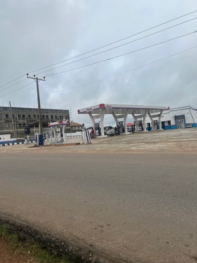 The image captures a cloudy day scene at a "Prevail" gas station in Ogbomosho, Nigeria. The gas station features a white canopy with red and blue trim, under which are several fuel pumps labeled "PMS 1," "PMS 2," "PMS 3," and "PMS 4." A white building with blue trim is attached to the canopy. In the foreground, a paved road leads towards the gas station, with a grassy and dirt shoulder on the left.
To the left of the gas station, a multi-story building is under construction, covered in scaffolding and dark netting. Several people are visible on the gas station premises. Two individuals appear to be standing near the building under construction. At the gas pumps, a silver car is being refueled, with at least two people standing nearby. Another person is seen near a motorbike on the right side of the gas station. A utility pole with multiple wires stands prominently in the middle left of the image. The overall environment suggests a commercial area with ongoing development.