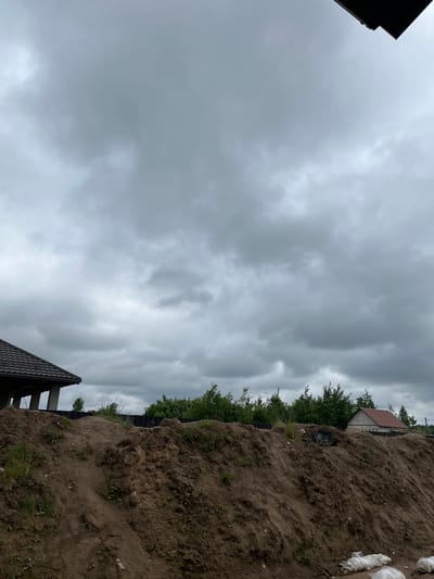 The image depicts a cloudy sky over a construction site. In the foreground, a large mound of earth, likely excavated during construction, dominates the lower portion of the frame. Scattered green vegetation, including grass and small plants, grows on the mound. In the middle ground, a row of trees forms a dense line. Beyond the trees, two houses are visible. The house on the left has a dark, tiled roof and is partially obscured. The house on the right has a red-tiled roof and appears to be constructed of light-colored materials. In the lower right corner, several white sacks are visible, possibly containing building materials or soil. The overall atmosphere is overcast and somber, suggesting the possibility of rain. The scene is situated in the Baravliany village council, Belarus.