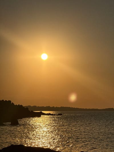 This is a vertical, outdoor, medium-shot photograph of the Black Sea coast at sunset, likely near Sozopol, Bulgaria. The sun, a bright orb of golden light, is positioned high in the orange-tinted sky, casting a shimmering path of light across the rippling water. In the foreground, dark, jagged rocks, possibly a breakwater or natural formations, line the left side of the frame, with the choppy water lapping against them. Silhouetted against the horizon is a faint, low-lying coastline with the suggestion of structures. The overall mood is serene and evokes the feeling of a tranquil evening by the sea. There are no people or animals visible in the image.