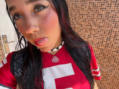 A young woman, with long, dark, wet hair with red highlights, poses for a selfie in front of a tiled wall. She is wearing a red Ohio State Buckeyes jersey, a black backpack, and a necklace made of shells. Her tongue is sticking out, and she has bold makeup on, including dramatic eyeliner and bright blush. A silver door handle is visible on the left side of the image. The background is a wall with small, square brown and tan tiles. The scene appears to be outdoors, possibly in the city of Pampatar, Venezuela.