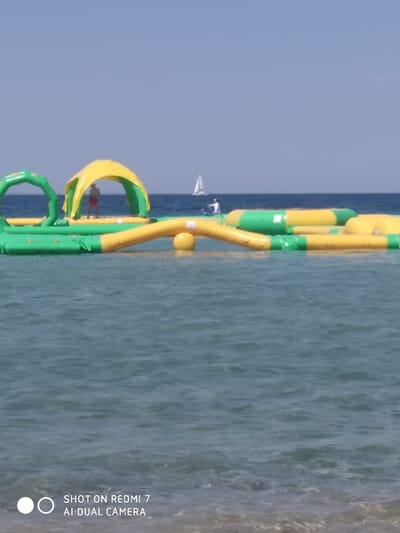 The image shows an inflatable water park on a clear, sunny day in the sea. The water park consists of green and yellow inflatable structures, including arches and a covered platform with a yellow canopy. A person is standing on the platform, wearing red shorts and a hat. In the distance, a sailboat can be seen on the calm blue water, under a bright blue sky. The setting appears to be a beach or coastal area, with the gentle waves of the sea in the foreground. The overall mood is one of leisure and outdoor recreation. The bottom left of the image contains text that reads "SHOT ON REDMI 7 AI DUAL CAMERA".