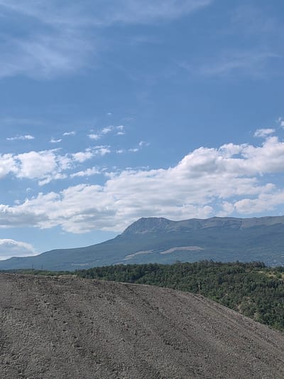 This media file depicts a landscape scene with a foreground of a large pile of dark-colored gravel or debris. Beyond the gravel pile, a dense, dark green forest covers rolling hills. In the background, a majestic mountain range stretches across the horizon, its peaks rugged and silhouetted against the sky. The sky is a vibrant blue, dotted with scattered white cumulus clouds that are illuminated by sunlight. The overall impression is one of a natural, somewhat wild environment. The lighting suggests it is daytime, likely late morning or early afternoon, with clear weather. There are no people or identifiable objects in the scene besides the natural elements and the gravel pile. The location appears to be rural or mountainous, consistent with the landscape cues. There is a small, light-colored cross or marker visible on the far right of the gravel pile, near the edge of the forest. There is also a faint, possibly man-made structure visible in the distance on the far left of the image, appearing as a vertical line against the distant hills. No text is visible in the image.