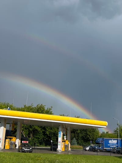 The image depicts a gas station with a prominent yellow canopy, featuring gas pumps. A double rainbow is visible against a cloudy, grey sky, suggesting recent or ongoing rain. To the right of the gas station, a blue truck with the word "LEADER" in white lettering on its side is parked. Further back, a black car is parked near the gas pumps, and a light-colored car is visible behind the truck. Lush green trees and grass surround the scene, indicating a rural or suburban setting. The lighting suggests it is daytime, possibly late afternoon or early evening, given the subdued light and cloudy conditions. The presence of a rainbow implies a break in the rain or clearing skies. The visible text on the truck reads "LEADER". The location context provided is Hlevakha, Ukraine.