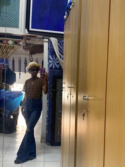 An individual is depicted taking a mirror selfie indoors. The subject, appearing to be female, is positioned centrally in the reflection, wearing sunglasses, a light-colored curly hat or wig, a short-sleeved brown top with a repeating geometric pattern, dark blue ripped flare jeans, and dark shoes. A golden necklace with a square pendant is visible. The subject holds a purple smartphone in their right hand, angled towards the mirror.
The reflected background reveals a diverse interior. To the reflected subject's left, a voluminous blue object, possibly fabric or a bag, rests on a dark surface. Behind the subject, multiple dark-colored items, possibly bags or keychains, are suspended against a light-colored wall. Above, various framed artworks or textile pieces are displayed, including a prominent blue abstract painting and other patterned designs. A section of the wall to the reflected subject's right features blue and white patterned tiling or fabric with a starburst motif. The floor is light-colored and reflective.
The right side of the image, not part of the reflection, shows a series of vertically oriented light-colored wooden cabinets or lockers. Each cabinet features a chrome handle with a key inserted into its lock. A blue textile item is partially visible atop the cabinets in the upper right corner.
The setting suggests a commercial space, such as a boutique, showroom, or a retail store, possibly incorporating a changing room or storage area. The specific location in the city of Alaguntan, Nigeria, cannot be confirmed from visual evidence within the image.