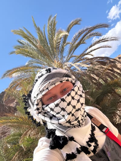 A person wearing a black and white keffiyeh stands in front of a palm tree under a blue sky with a few white clouds. The keffiyeh covers most of their face, leaving only their eyes visible. They are wearing a white shirt and a gray vest with a red and black strap. The location is Dahab, Egypt, as indicated by the vegetation and attire.