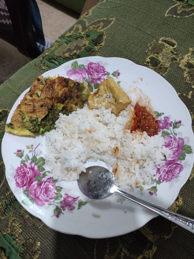 A white plate with a floral design sits on a green and gold tablecloth. On the plate are a serving of white rice, a piece of fried tofu, a slice of omelet with green vegetables, and a small amount of red chili sauce. A metal spoon lies on the plate to the right of the rice. The scene is located in the city of Kecamatan Lawang, Indonesia, at coordinates (latitude: -7.828, longitude: 112.701). There are no people in the image.