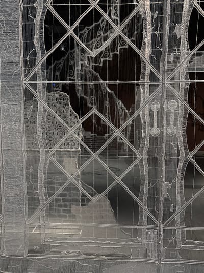 The image displays a translucent, white, 3D-printed architectural screen or facade, intricately designed to resemble a traditional window or ornate grille. The screen occupies the majority of the foreground, featuring a grid of intersecting diagonal lines forming diamond-shaped panes, interspersed with vertical, architecturally-inspired elements that mimic pilasters or mullions. These vertical sections exhibit a textured, almost corrugated appearance, and on the right, two distinct knob-like handle forms are present. The construction of the screen reveals its filament nature, with visible fine strands and layered patterns indicative of 3D printing technology.
Through the translucent material, a dimly lit, out-of-focus background is discernible. On the left side, an abstract, textured, and porous sculptural form is visible. Towards the center-right and bottom, sections of a reddish-brown brick wall, characteristic of older European architecture, can be seen. The overall background suggests an interior space, possibly a historical building or courtyard.
This specific type of delicate, 3D-printed architectural installation, often evoking Venetian lace and Gothic design elements, is associated with contemporary art and architecture exhibitions, such as those featured during the Venice Architecture Biennale in Venice, Italy. These installations are frequently placed within the city's historic palazzi or exhibition spaces, creating a dialogue between modern technology and traditional Venetian aesthetics. No people, discernible subjects, or explicit actions are present within the frame; the scene focuses entirely on the interplay between the foreground installation and its architectural backdrop.