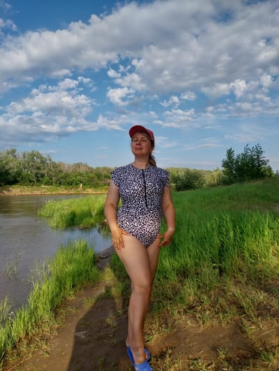 A woman in a leopard-print one-piece swimsuit and a red baseball cap stands on a dirt path near a river. She is facing forward with her eyes closed and her head tilted back slightly. Lush green grass and trees line the riverbank and extend into the background, where a cloudy blue sky is visible. The scene appears to be a natural outdoor setting, possibly a riverbank in the city of Chkalov, Russia.