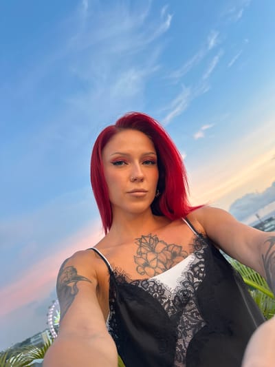 A medium shot depicts a fair-skinned woman with red hair, facing the camera. Her body is oriented towards the right side of the frame. She has a tattoo on her chest, and is wearing a black lace-trimmed top with white straps. The woman is positioned against a gradient sky transitioning from blue to orange. The location appears to be near Tibubeneng, Indonesia, as implied by visible palm fronds and architectural details. Tattoos adorn both her upper arms.