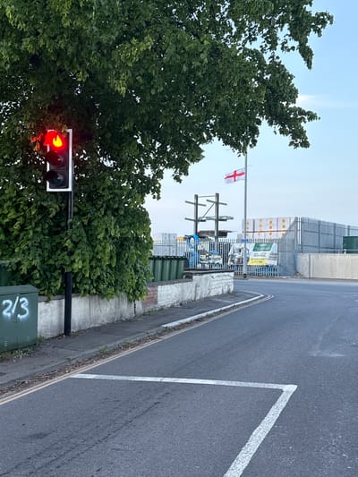The image depicts a street scene with a red traffic light, partially obscured by a tree, and a flag of England flying from a pole in the background. The road is asphalt with white painted lines marking a pedestrian crossing. A metal fence and stacked building materials are visible behind the flagpole. A concrete wall runs alongside the road, and a green electrical box with white graffiti is located near the traffic light. The scene appears to be in Great Yarmouth, United Kingdom based on the English flag.