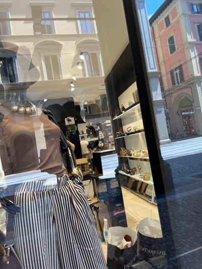 A daytime shot, heavily distorted due to reflections, shows a storefront, likely in Bologna, Italy. The window reflects a light-colored building across the street with ornate window frames. Inside the store, one can see jewelry displayed on shelves, along with clothing. A person is visible in the interior, standing near what appears to be a display or counter. The objects inside the store are obscured by reflections of the exterior building.