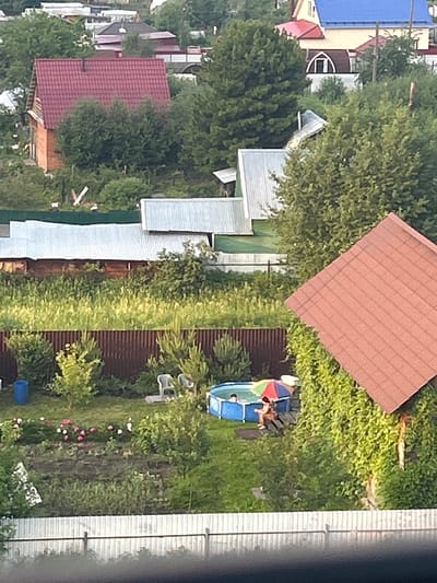 The image shows an aerial view of a backyard scene in Kemerovo, Russia. The scene is filled with lush green trees and vegetation, suggesting a warm, possibly summer day. There are several residential buildings with red and silver roofs, indicating a suburban or semi-rural setting.

In the center of the yard, a blue inflatable pool is set up. Two people are visible in the pool, one lying down and another sitting, enjoying the water. A colorful umbrella provides shade over the pool area. Adjacent to the pool, there are lounge chairs and other outdoor furniture. The yard also features a flower bed with pink and white flowers, adding to the vibrant atmosphere. A dark brown wooden fence runs along the side of the property.

The time of day appears to be late afternoon or early evening, with the sun casting soft light on the scene. The overall mood of the image is one of relaxation and leisure, with the individuals in the pool clearly enjoying themselves. There is no visible text in the image.