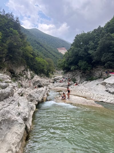 In the town of Cagli, Italy, a natural rocky gorge features a flowing river with clear, greenish water. Several people are present, some bathing or wading in the shallow parts of the river, while others are sunbathing or resting on the smooth, light-colored rock formations that line the gorge. A woman in a black swimsuit and a young boy in blue shorts are standing on a rock near the water's edge, with the boy sitting and the woman standing. The gorge is surrounded by lush green trees and steep, forested hills. The sky above is overcast with some patches of blue. The overall scene depicts a popular natural swimming spot.