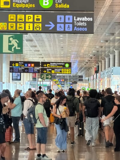 The image depicts a crowded interior scene at an airport terminal in El Prat de Llobregat, Spain. Overhead signs in English and Spanish provide directions for baggage claim, taxis, metro, and restrooms. Several people, dressed in casual summer attire, are walking in various directions, some with luggage. A large group of individuals are visible in the foreground, navigating the space. The environment is characterized by a polished floor reflecting the overhead lighting and architectural elements like columns. The overall atmosphere suggests a busy transit hub.