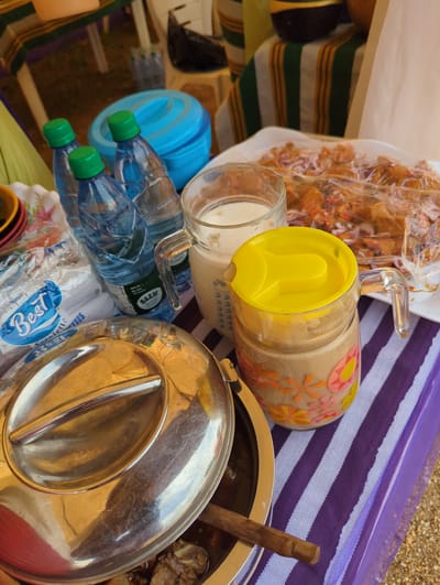 The image presents an outdoor scene with a table setup for a meal, likely in the city of Maigizo, Nigeria. The table is covered with a striped purple and white cloth. Several items are arranged on the table: a clear plastic bag containing fried food mixed with red onion and spices, a glass pitcher with a yellow lid and floral decorations, a clear glass cup, a blue water bottle with a green cap, a pack of "Best" brand serviettes, a stainless steel serving dish with a lid, and a wooden spoon inside the dish. The setting appears casual and utilitarian.