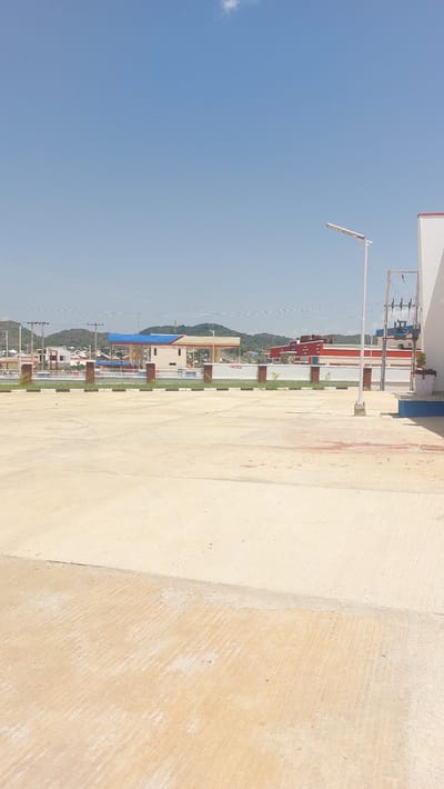 The image displays a bright, sunny day in Kuru Dungus, Nigeria. The scene is dominated by a large expanse of light-colored, textured concrete in the foreground, suggesting an outdoor paved area like a walkway, plaza, or open space. To the left, a row of colorful buildings, likely shops or offices, lines a street. These buildings feature a mix of white, red, and blue elements, with some appearing to be multiple stories high. A street lamp stands tall in front of the buildings, and utility poles with wires are visible, indicating a developed area. In the background, lush green hills rise, with more buildings scattered at their base and along the slopes, hinting at a town or city nestled within the landscape. The sky is a clear, vibrant blue, with a small wisp of a cloud. The overall impression is one of a well-lit, active urban or semi-urban environment on a pleasant day. No people are visible in the frame. There is no discernible text in the image.