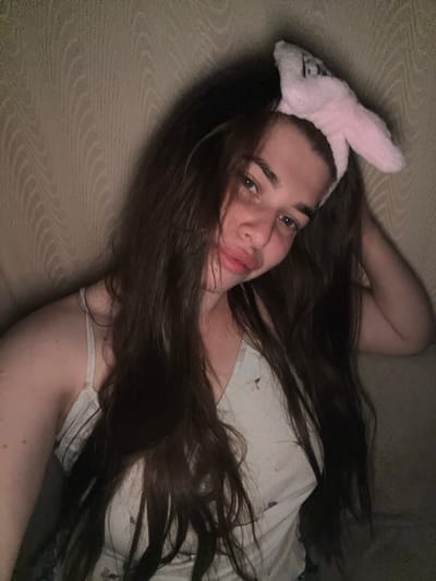 In this selfie taken in the city of Мещёры, Russia, a young person with long, dark brown hair is looking at the camera. They are wearing a light-colored tank top with a delicate pattern and a fluffy pink headband with bunny ears. The background appears to be a textured wall or fabric, possibly a couch or a wall covering, with a wavy pattern. The lighting is dim, creating shadows and highlighting the person's face and hair. The person's left hand is resting near their face, and their right arm is bent at the elbow, with their hand near their head. The overall impression is that of a casual, intimate moment captured in a domestic setting.