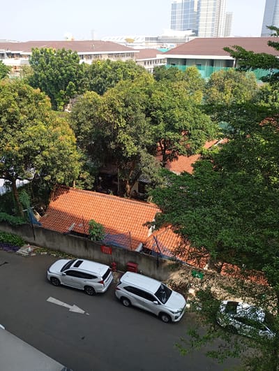 In an aerial view of a residential area in South Jakarta, Indonesia, two white SUVs are parked on a paved street. One SUV, a Mitsubishi Pajero Sport, is facing left, while another white SUV, possibly a Mitsubishi Xpander, is parked behind it, facing right. The street has a white arrow marking indicating traffic direction. Behind the street, a grey concrete wall separates it from a property with red-tiled roofs and lush green trees. In the background, modern high-rise buildings are visible against a hazy sky. The scene appears to be a typical urban residential setting in the morning light.