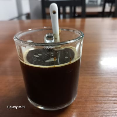 A close-up shot shows a glass of dark liquid, likely coffee, on a wooden table. A white spoon is partially submerged in the liquid, with a small hole visible in its handle. The rim of the glass has a frothy residue. The background is blurred, showing dark shapes that could be chairs and a bright white area that might be a wall or window. A watermark in the bottom left corner reads "Galaxy M32." The scene is devoid of people and specific landmarks, making it impossible to determine its location in Olsztyn, Poland, or any other city.