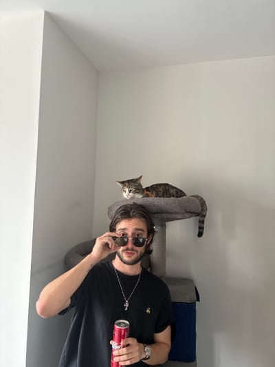 A man and a cat are situated in an indoor setting, potentially in a residence in Huixquilucan de Degollado, Mexico. The man, wearing a black t-shirt, sunglasses, and a silver necklace, holds a red Dr. Pepper can in his right hand and adjusts his sunglasses with his left. He has dark hair and a beard. To the left of the man is a cat, resting on top of a cat tree/scratching post. The cat is tabby-colored. Behind the man and the cat are cushions or pillows, possibly on a couch. The background is a white wall with some visible molding or corners. The image is rotated.