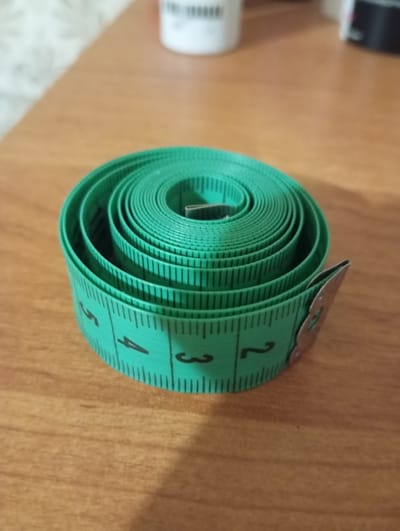 The image displays a coiled green measuring tape on a wooden surface. The measuring tape has black markings and numbers, with visible numbers 2, 3, and 4. The background is blurred, showing a light-colored wall and some out-of-focus objects, one of which appears to be a white cylindrical container with black text. There is no indication of the location being Orenburg, Russia, or any city for that matter. No people or actions are depicted in the image.