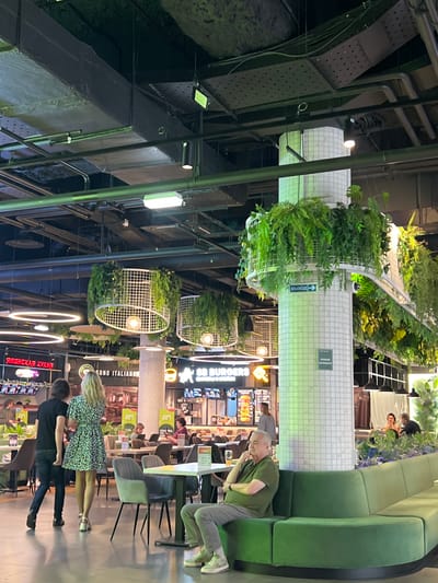 This image depicts an interior scene, possibly a food court or a restaurant area within a large building, given the presence of multiple dining establishments and seating arrangements. The environment appears to be a modern indoor space with a high ceiling and industrial-style exposed infrastructure, including pipes and ventilation systems. Decorative elements include hanging plants and circular lighting fixtures, creating a blend of natural and contemporary aesthetics.

Several people are visible in the scene, engaged in various activities. In the foreground, a man in a green t-shirt and grey pants is seated, looking down. Behind him, a couple is standing, with the woman wearing a patterned dress and the man in a dark shirt. Further in the background, other individuals are seen dining or walking. The lighting suggests it is likely evening or indoors with artificial illumination.

Visible text includes signs for "BURGER" and "ЯПОНСКАЯ КУХНЯ" (Japanese Cuisine) in Russian, and "SONO ITALIANO" (possibly meaning "It's Italian" or "Sono Italiano Restaurant") in English and Italian. There is also a sign with an arrow and the Russian word "ВЫХОД," meaning "Exit." These cues indicate a diverse food court or restaurant setting. The overall mood appears casual and social, typical of a public dining area. The location context of Ladozhskoe Ozero, Russia, further supports the interpretation of a Russian establishment.