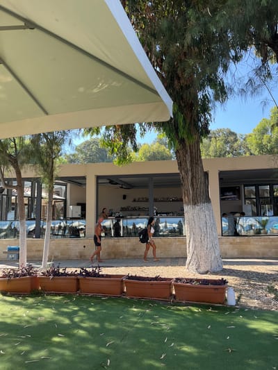 The image depicts an outdoor scene in Germasogeia Municipality, Cyprus, likely a beachfront establishment. In the foreground, a large, light-colored umbrella casts shadows on artificial turf. A row of rectangular planters filled with dark-leaved plants separates the turf from a paved walkway.
On the walkway, two individuals are walking. A man in black shorts is walking to the left, and a woman in a white top and patterned shorts, carrying a black backpack, is walking to the right.
Behind them is a building with a modern facade, featuring large glass windows and an open-air bar area. Bottles are displayed on shelves behind the bar, where a woman in a black top is visible. Sunlight filters through trees, casting dappled shadows on the scene. The background shows lush green trees and a clear blue sky, suggesting a warm, sunny day. The overall atmosphere appears relaxed and summery.