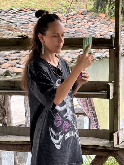 A woman is holding a green smartphone in her left hand, appearing to take a photo or video. She is wearing a dark grey t-shirt with a skeleton and butterfly graphic, and has a ponytail. A pair of sunglasses hangs from her shirt. She is wearing silver earrings. She is standing in a wooden structure. The background consists of tiled roofs and some vegetation. The location is Lũng Cú Commune, Vietnam, evidenced by the traditional architecture.