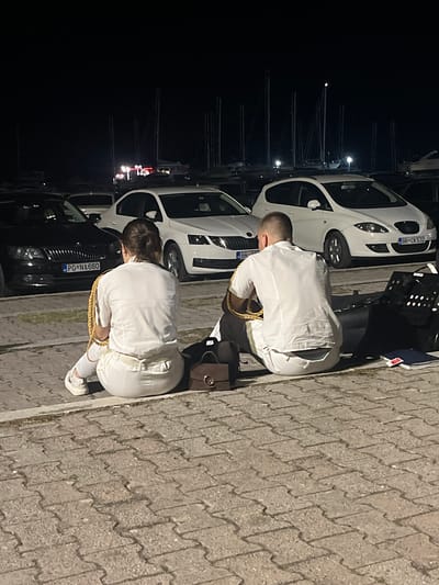 In Šušanj, Montenegro, two individuals, a male and a female, are seated on a paved surface at night. They are facing away from the camera, dressed in matching white uniforms with gold trim and epaulets. The male is on the right and the female is on the left. They appear to be wearing similar white pants and shoes. The female has her hair tied back in a bun.

Behind them, several white cars are parked, with license plates visible on two of them. The license plate on the black car in the foreground on the left reads "PG NA 660". The license plate on the white car in the middle reads "BR CK 532". In the background, numerous sailboat masts are visible against a dark sky. The scene suggests a harbor or marina setting.

To the right of the male, a black object with a grid pattern is visible, resembling a music stand or a similar piece of equipment. A brown handbag is placed on the ground between the two individuals. The lighting suggests artificial illumination from nearby sources.