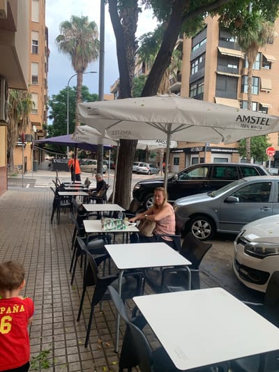 The image depicts an outdoor seating area of a cafe or restaurant located on a street in Valencia, Spain. The scene is set on a tiled sidewalk with several tables and chairs. In the foreground, a young boy in a red shirt with "España 6" on the back is walking away from the camera. Behind him, a woman with blonde hair is sitting at one of the tables, looking towards the camera. She is wearing a pink top and carrying a beige handbag. Further down the sidewalk, a man in an orange shirt is standing near a purple umbrella, and another man in a black shirt is seated at a table. Several cars are parked along the street, including a black SUV and a grey sedan. The cafe's umbrellas are branded with "AMSTEL 100% MALTA." The buildings in the background are typical of urban architecture in Spain, with balconies and awnings. Palm trees are visible, suggesting a Mediterranean climate. The overall impression is a casual outdoor dining environment on a city street.
