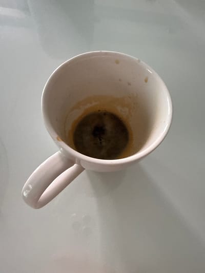 A light pink ceramic mug, with remnants of dark liquid and a thin film of residue coating the interior, rests on a light gray, reflective surface. The mug is positioned at an angle, with its handle extending to the left. The interior of the mug shows a concentrated dark substance at the bottom, with lighter brown residue extending up the sides. The surrounding surface appears to be a tabletop or counter, exhibiting subtle reflections and soft shadows. There are no people or other objects visible in the immediate vicinity. The image does not contain any contextual information that would suggest a specific location in the city of Gabrovo, Bulgaria.