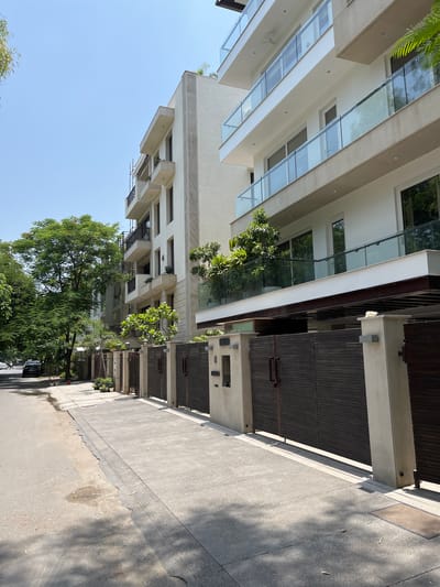 The image shows a street scene in New Delhi, India, likely a residential area. On the right side of the frame, there are modern multi-story buildings with white facades, balconies with glass railings, and dark wooden gates. The buildings appear to be upscale residences. A paved sidewalk runs in front of the gates, bordered by the street on the left. Lush green trees line the street and are visible in front of and between the buildings, providing shade and a natural element to the urban environment. The sky is clear and blue, indicating a sunny day. A few cars are visible parked or driving on the street in the background. The overall impression is of a well-maintained, affluent neighborhood in New Delhi.