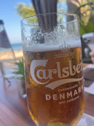 The image is a close-up shot of a glass of Carlsberg beer, condensation visible on the exterior, placed on a wooden table outdoors. In the background, blurred elements suggest a coastal setting with a body of water and possibly palm trees, consistent with a location like Protaras, Cyprus. The beer glass displays the Carlsberg logo, the royal Danish crown, "BY APPOINTMENT TO THE ROYAL DANISH COURT", "COPENHAGEN DENMARK", and "1847 ONWARDS". A bottle of water is visible to the left, and a hand holding a mobile phone is partially seen on the right. The lighting indicates it is daytime, possibly late afternoon due to the warm tones.