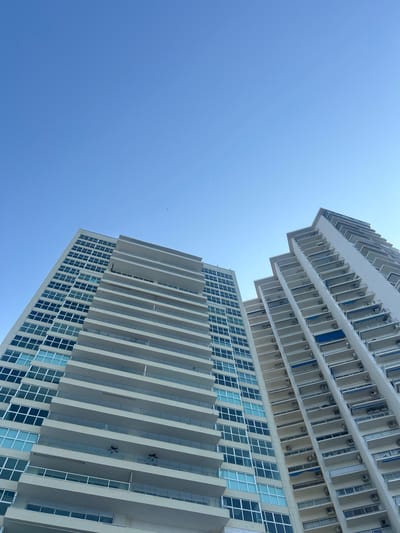 The image, taken from a low angle, features two tall buildings against a clear, bright blue sky. The lower building has a modern design with rows of windows and white concrete balconies. The upper building has a similar architectural style with balconies and windows and rows of AC units. The buildings are located in Acapulco, Mexico.