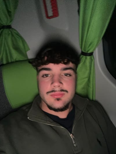 A young man with dark curly hair and a trimmed beard lies sideways in what appears to be an upper berth of a bus or train. He looks towards the camera with a serious expression. He is wearing a dark shirt and a green jacket. Behind him, a green curtain is tied back with a black band. The walls and the headrest seem to be gray or white, with a red object visible in the background, possibly a luggage rack.
