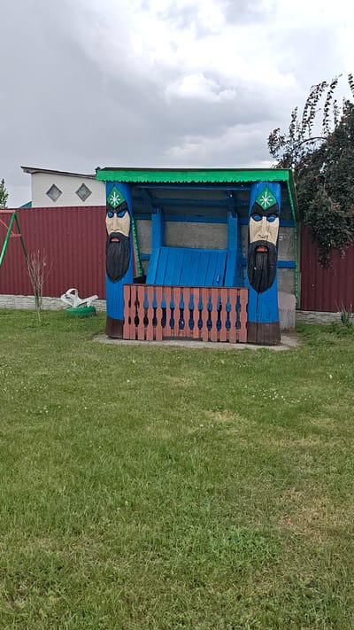 A wooden gazebo with a green roof and blue interior is situated in a grassy yard. The gazebo is decorated with two carved wooden faces on its front posts, each wearing a green headdress with a white asterisk. The gazebo's front railing is made of reddish-brown wooden slats. In the background, a red corrugated metal fence runs horizontally, with a white building with diamond-shaped windows behind it. A swingset with green and red elements is partially visible to the left of the gazebo. The ground is covered in green grass, with some patches of dry grass and scattered white flowers. The sky is overcast with gray clouds. The image appears to have been taken in the city of Revne, Ukraine.