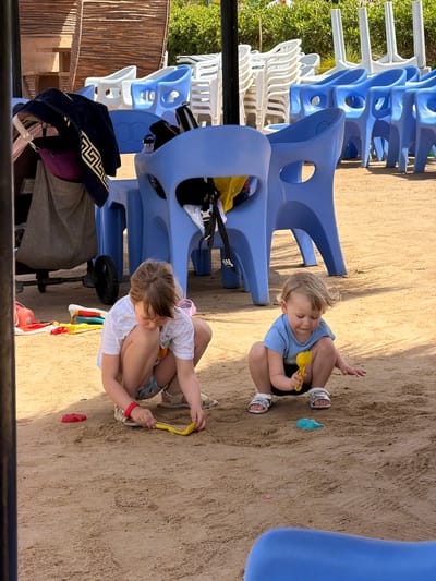 Two young children are playing in a sandy area. One child is fair-haired and wearing a light blue shirt and black shorts with white sandals. The other child is wearing a white shirt and floral patterned shorts with tan sandals. The children are positioned near several stacked, blue plastic chairs. In the background, there are additional stacks of white and blue plastic chairs, as well as some greenery and a wooden fence. A stroller and a beach bag with a black hat on top are also visible. The sandy area appears to be a beach or recreation area possibly in Hurghada, Egypt.