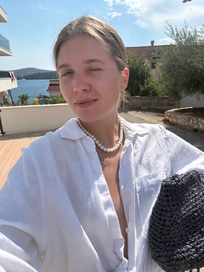 The media file shows a young woman from the chest up, wearing a white linen shirt and a pearl necklace with a cross pendant. She has blonde hair pulled back and is looking towards the camera with a slight smile and one eye closed, as if winking. A dark woven bag is visible over her shoulder.
The background suggests an outdoor setting with a view of the Adriatic Sea and a coastline with buildings. The sky is blue with some clouds, indicating it is daytime. The lighting suggests it is a sunny day, likely late morning or early afternoon. The architecture of the buildings in the background and the presence of the sea suggest a Mediterranean location, consistent with the provided location context of Grad Šibenik, Croatia. The overall mood is relaxed and cheerful, possibly a vacation or a day out enjoying the weather. No text is visible in the media file.