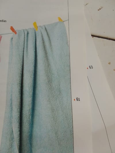 The image shows a close-up of a light blue towel draped over a clothesline, secured by two yellow and two orange clothespins. The towel is wrinkled, creating vertical folds. To the right of the towel, a white sheet of paper is partially visible, with two numerical annotations, "63" and "61", each preceded by a small red triangle. The paper appears to be a diagram or instruction sheet. The background to the left of the towel contains the word "edias" in dark text. The scene is an indoor or outdoor setting where laundry is being hung to dry. There is no indication of the specific location being in the city of La Tagua, Venezuela.