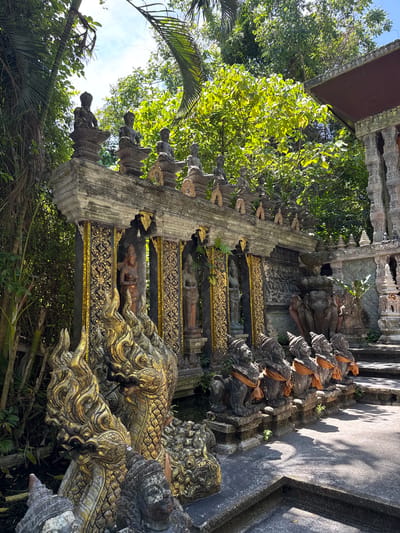 The image depicts an outdoor shrine in Baan Lipa Noi, Thailand. The shrine features ornate stone sculptures of mythological creatures and deities, with a backdrop of lush green foliage and trees. In the foreground, a series of intricately carved golden serpent-like statues with multiple heads are visible. Behind them, a row of seated lion-like statues, adorned with orange sashes, stand guard. The main structure of the shrine is a long, tiered stone edifice with niches containing statues of human-like figures, possibly deities or historical figures. These niches are decorated with gold accents and intricate patterns. The overall atmosphere is one of ancient artistry and spiritual significance, set within a tropical environment.