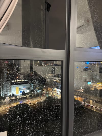 The image captures a nighttime view of a city skyline from a window covered in raindrops. The scene depicts a busy urban environment, likely Kuala Lumpur, Malaysia, given the architectural style and density of buildings. Through the rain-streaked glass, illuminated buildings and a highway with moving vehicles are visible, suggesting a vibrant, active city. The foreground is dominated by the window frame and the water droplets, which create a diffused, atmospheric effect on the cityscape beyond. The lighting suggests artificial sources, typical of a city at night, with streaks of light from car headlights and building illuminations. The weather condition is clearly rainy, indicated by the numerous water droplets on the glass. The overall impression is one of a bustling metropolis viewed during a downpour.