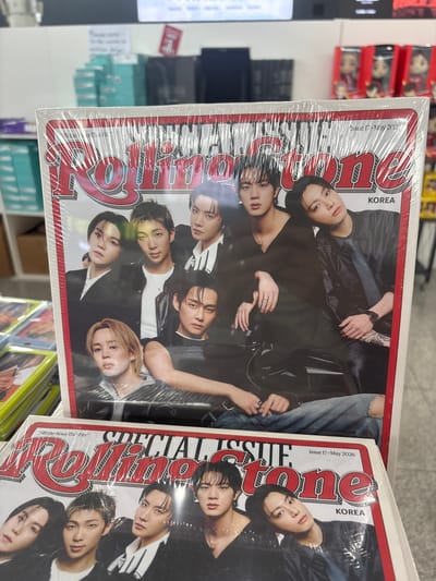 The image shows two copies of Rolling Stone magazine stacked on top of each other, with the top copy partially obscuring the bottom one. Both magazines feature the same cover image, which prominently displays seven individuals, likely members of the K-pop group BTS, dressed in dark clothing. The magazine title "Rolling Stone" is in large red letters across the top of both covers. The top magazine has text that reads "Issue 17 · May 2026" and "KOREA" in smaller print. The bottom magazine also has text that reads "SPECIAL ISSUE" and "Issue 17 · May 2026" and "KOREA." The magazines appear to be shrink-wrapped. The background suggests an indoor retail environment, possibly a bookstore or convenience store, with shelves of merchandise visible. The lighting is bright and artificial. The image is taken from a slightly elevated angle, looking down at the magazines. There are no explicit visual cues to pinpoint the location within the city of Busan, South Korea.
