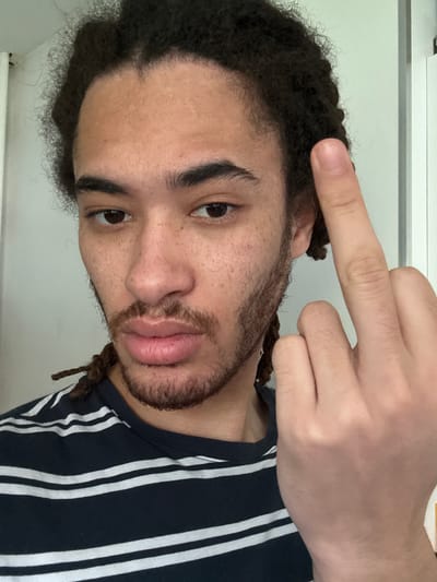 A close-up, medium shot depicts a light-skinned male with dark curly hair and a partial beard. He is wearing a navy blue and white striped shirt. His left hand is raised, showing only the middle finger extended. The background is a light-colored wall. The image was not taken in Les Ulis, France.
