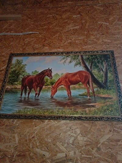 The image displays a framed painting mounted on a textured wall composed of Oriented Strand Board (OSB) or chipboard, situated in Gorka, Russia. The painting is oriented diagonally, with its lower-right quadrant closer to the viewer. It depicts two horses in a body of water, likely a river or stream, under a blue sky with some clouds.
The horse on the left is dark brown/bay, standing in the water with its head slightly raised and facing right, its front legs submerged to just below the chest. The horse on the right is a lighter reddish-brown/chestnut, positioned with its head lowered to the water, actively drinking. Its front legs are also submerged, with water reaching approximately its knees. The water reflects the sky and portions of the horses. The background within the painting shows green trees and foliage along the water's edge. The painting is encased in an ornate frame, dark with gold-colored decorative scrollwork. Above the painting, a white electrical cable is visible, secured horizontally across the OSB wall by white clips or ties.