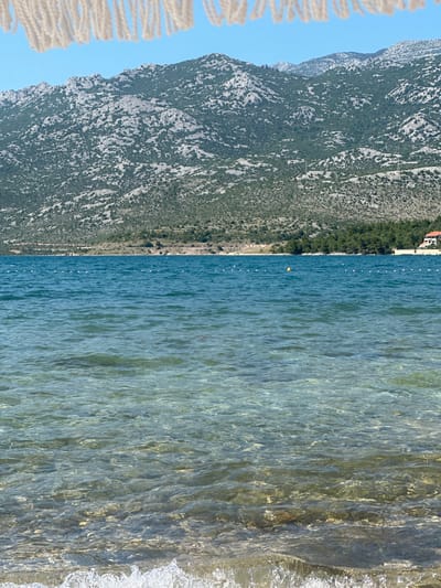 A vertical, eye-level shot captures a serene coastal scene in Rovanjska, Croatia. In the foreground, clear, shallow water laps at a sandy shore, with the sandy seabed visible beneath the translucent waves. The water transitions to a deeper, more opaque blue further out, marked by a line of white buoys. In the background, a densely forested hillside slopes upwards to meet rugged, rocky mountains under a clear blue sky. A single red-roofed building is visible amidst the trees on the right side of the frame. A fringe of white tassels, possibly from an umbrella or canopy, hangs from the top edge of the image, framing the view. The scene is devoid of people.