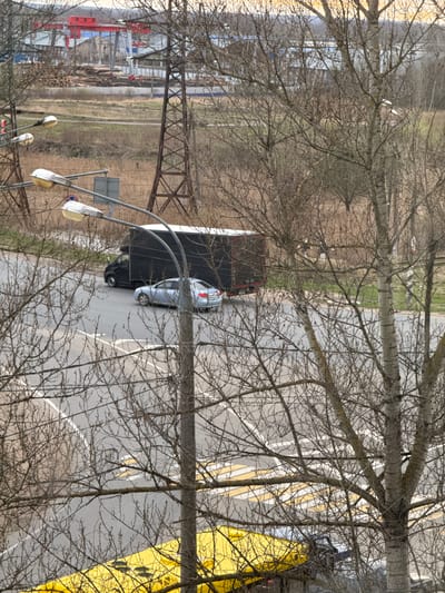The image presents an elevated view overlooking a road, bare trees, and an industrial or construction area, likely during late autumn or early spring due to the leafless trees. The scene is lit by diffused daylight, suggesting an overcast sky or hazy conditions.
In the foreground, a dense network of bare tree branches frames the view. Below these branches, a paved road stretches across the middle ground. On the road, a dark-colored truck, possibly a large delivery vehicle or a semi-truck cab, is partially visible, followed by a light-colored sedan (silver or light blue). Road markings, including solid white lines and diagonal yellow lines, are discernible. To the right of the road, a prominent, bright yellow structure or banner with dark markings, possibly a biohazard symbol, takes up a significant portion of the frame. Streetlights with multiple lamps are visible on the left side of the road.
Beyond the road and trees, a wide, open field with dry grass extends into the distance. In the far left-middle ground, a large, dark brown metal truss structure, possibly part of a bridge or industrial gantry, spans across the landscape. Further left, in the background, red and blue industrial equipment, resembling cranes or parts of a factory, are visible, alongside piles of what appears to be construction material or debris.
No people are explicitly visible, but the vehicles on the road imply human presence and activity. The overall setting combines elements of urban infrastructure, natural landscape, and industrial development, consistent with an area like Novonikolskoye, Russia. No clear, readable text is present, aside from the ambiguous markings on the yellow structure and a partially visible circular traffic sign with a blue background and red border.