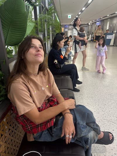 The image depicts a group of people in what appears to be an airport waiting area. In the foreground, a young woman with long brown hair and blue eyes is looking upwards, with a beige t-shirt adorned with colorful embroidered patches. A red and black plaid shirt is draped around her shoulders. Her left arm rests on her lap, and her right arm is crossed over her chest. To her right, a person wearing a denim jacket and jeans is seated, with one foot in a black sandal visible.

In the background, several other individuals are visible, including a woman carrying a baby in a carrier, a child in a purple t-shirt and pink pants, and other people sitting and standing. The setting features a speckled floor and what looks like modern airport seating. The lighting suggests it is daytime. One of the background elements includes a sign with Korean text, likely indicating airport information. The overall scene conveys a sense of waiting, common in travel settings, with a relaxed atmosphere.